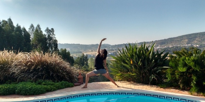 lady in yoga pose reverse warrior outside by a pool with a backdrop of hills in a European country in the summer with green plants and trees around her
