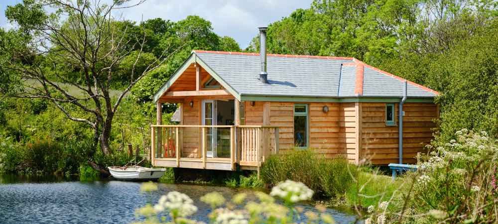 Image of lakeside wooden cabin in the sunshine with flowers in the foreground and woodland in the background. Peaceful yoga retreat in Cornwall
