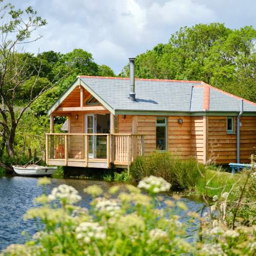 Image of lakeside wooden cabin in the sunshine with flowers in the foreground and woodland in the background at a peaceful yoga retreat in Cornwall.