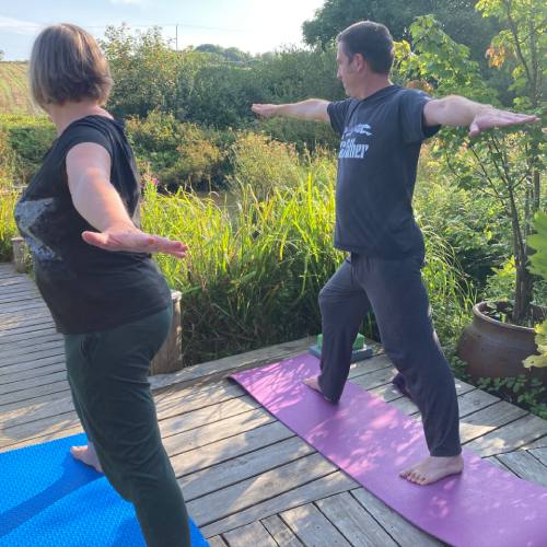 Man and woman practising yoga in warrior 2 position looking out over the lake at the peaceful yoga retreat in Cornwall at Pengelly Retreat
